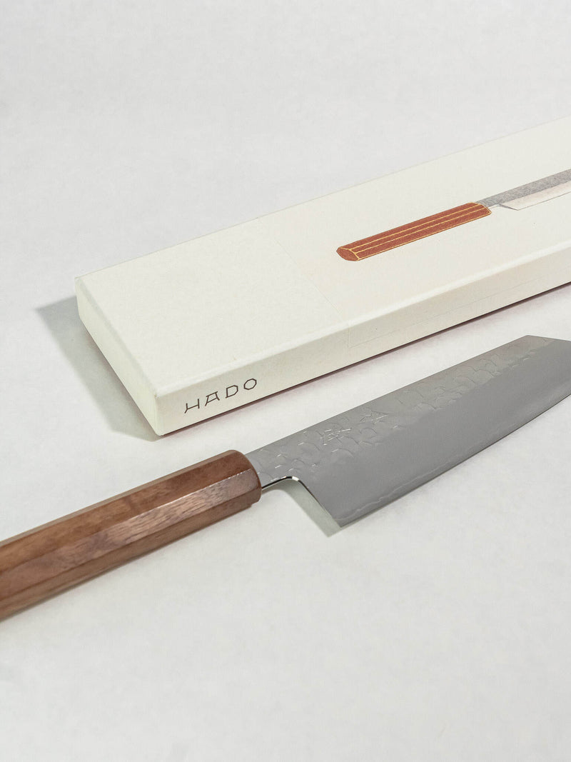A HADO Japanese kitchen knife with a dark wood handle and textured steel blade, shown next to its cream-colored presentation box on a light gray background.