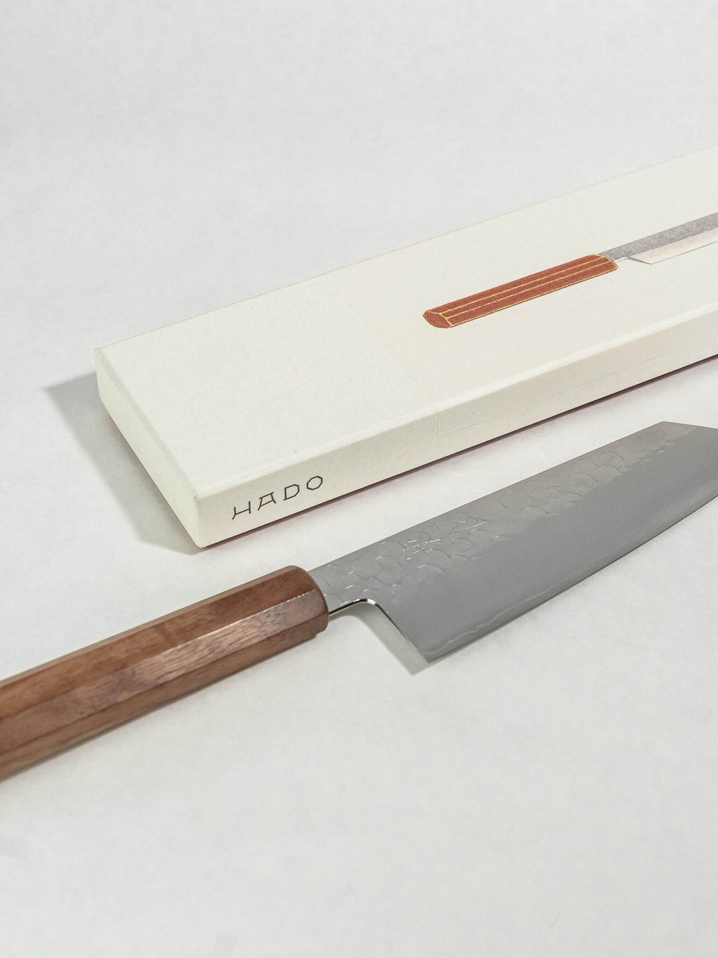 A HADO Japanese kitchen knife with a dark wood handle and textured steel blade, shown next to its cream-colored presentation box on a light gray background.