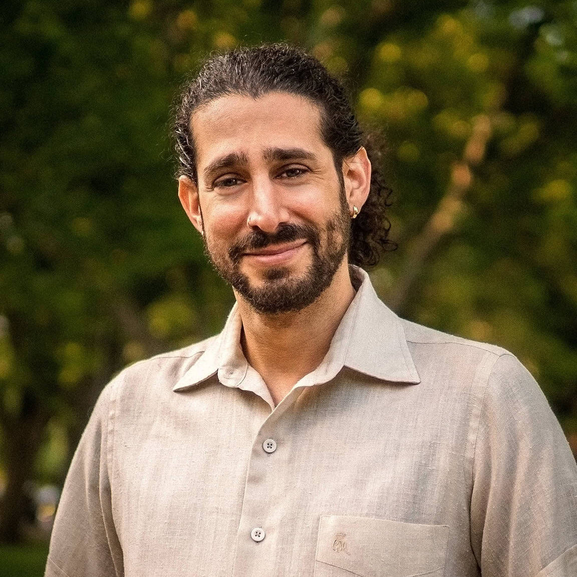 Man with a beard wearing a light-colored shirt outdoors with greenery in the background