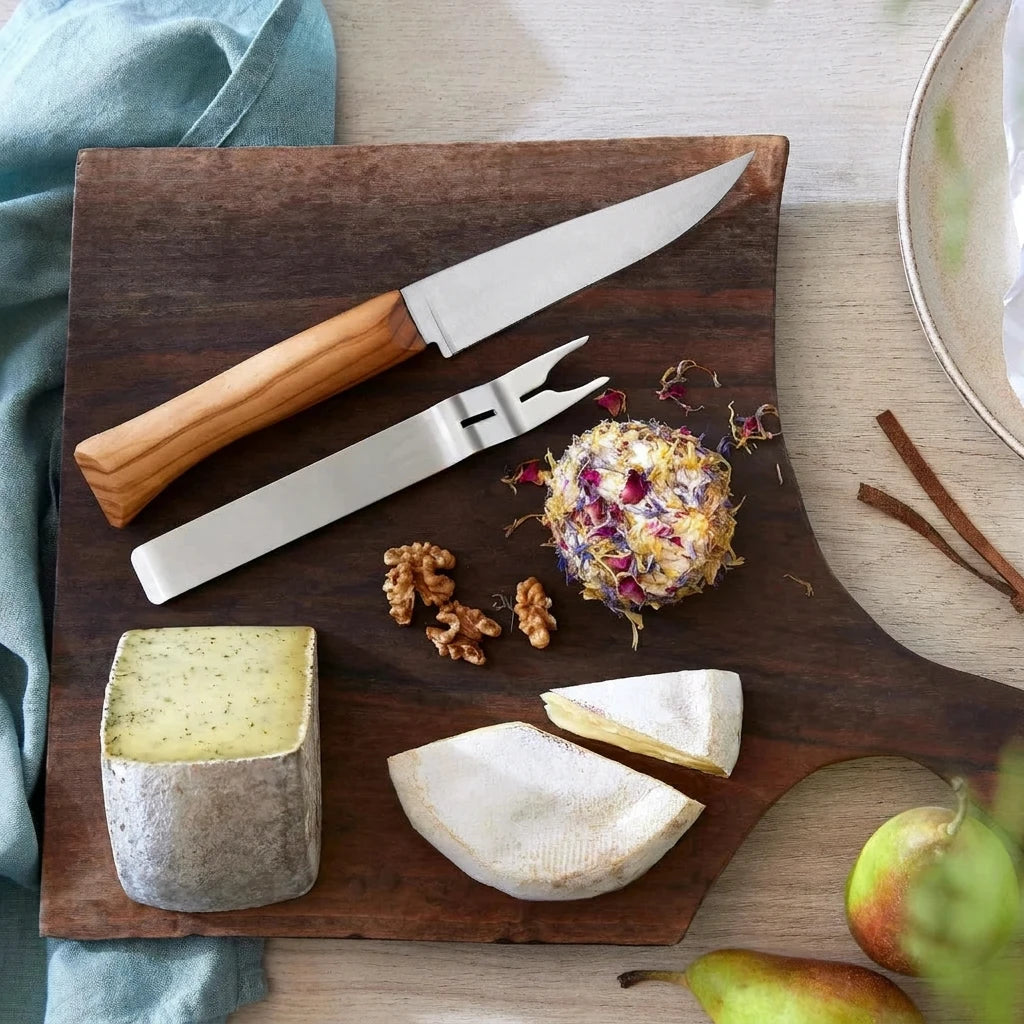 A wooden serving board styled with a stainless steel cheese knife, a fork, wedges of white cheese, a block of herb cheese, walnuts, dried flowers, cinnamon sticks, and pears on a light wood table.