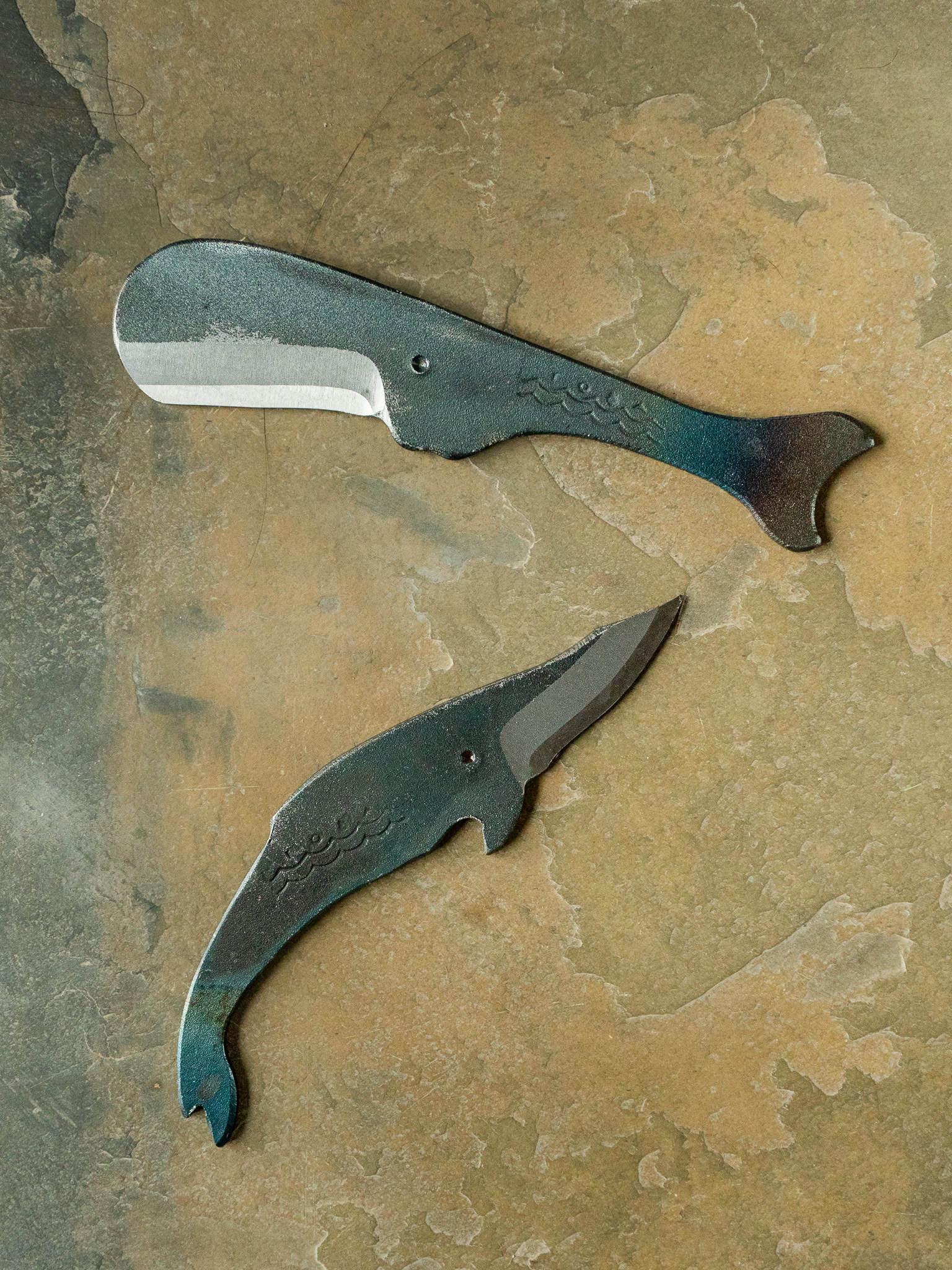 Two metallic whale shaped utility knives on a textured stone surface